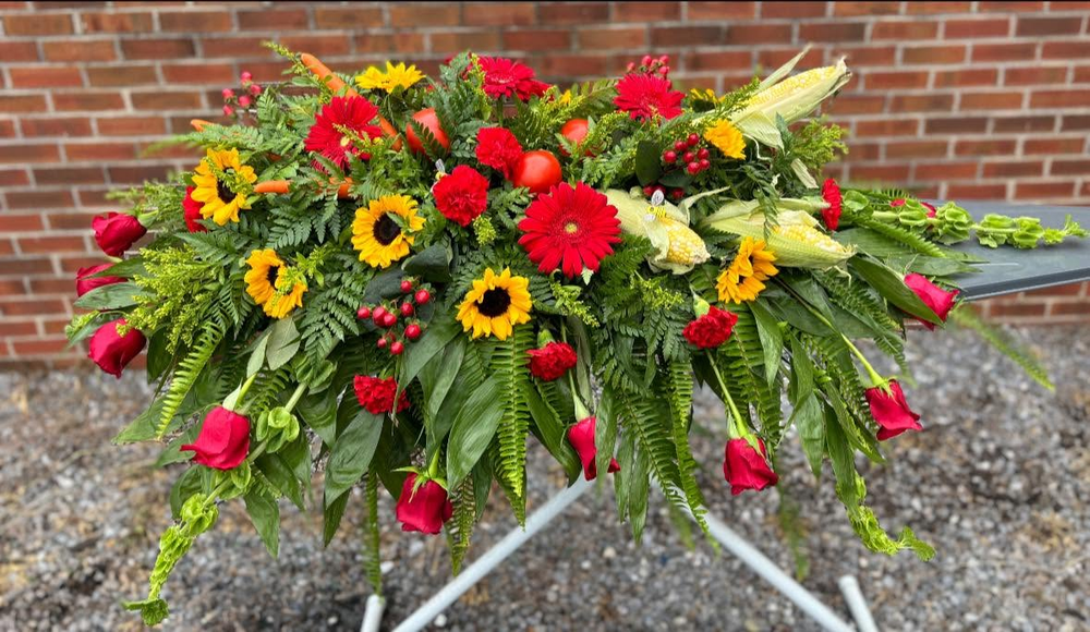 Casket spray of sunflowers, red gerbera daisies, roses, and garden vegetables